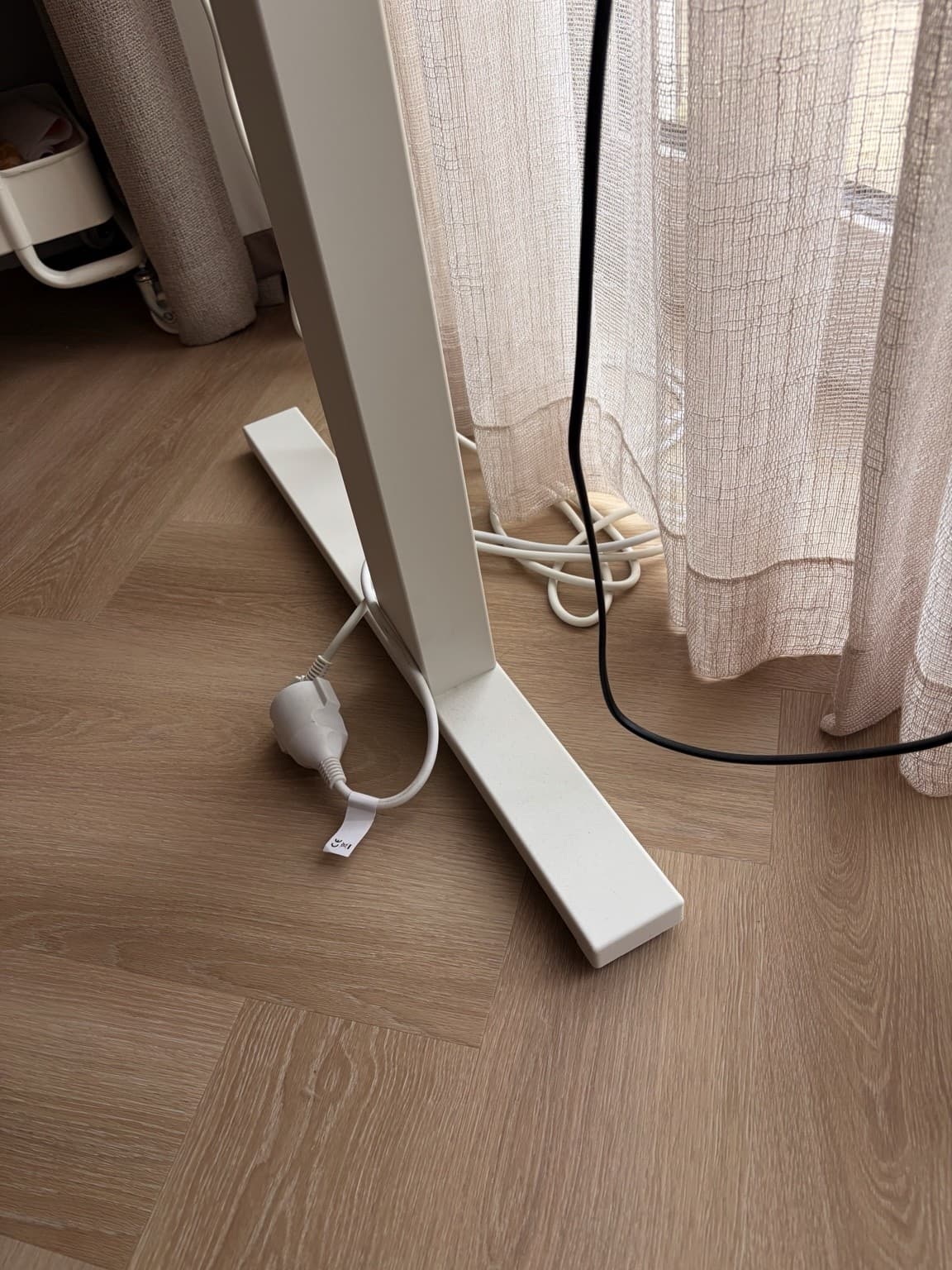 An extension lead and cables beside a white desk leg and sheer curtains.