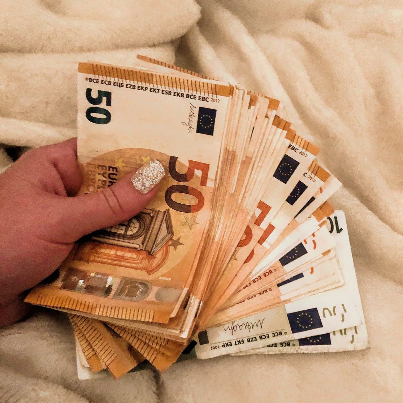 Euro banknotes fanned out in someone’s hand, resting on a cream blanket.