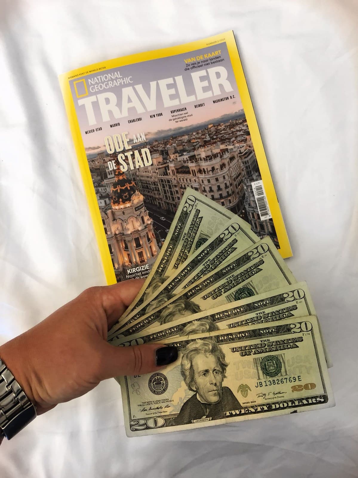 US dollar bills fanned out next to a National Geographic Traveler magazine and a watch.
