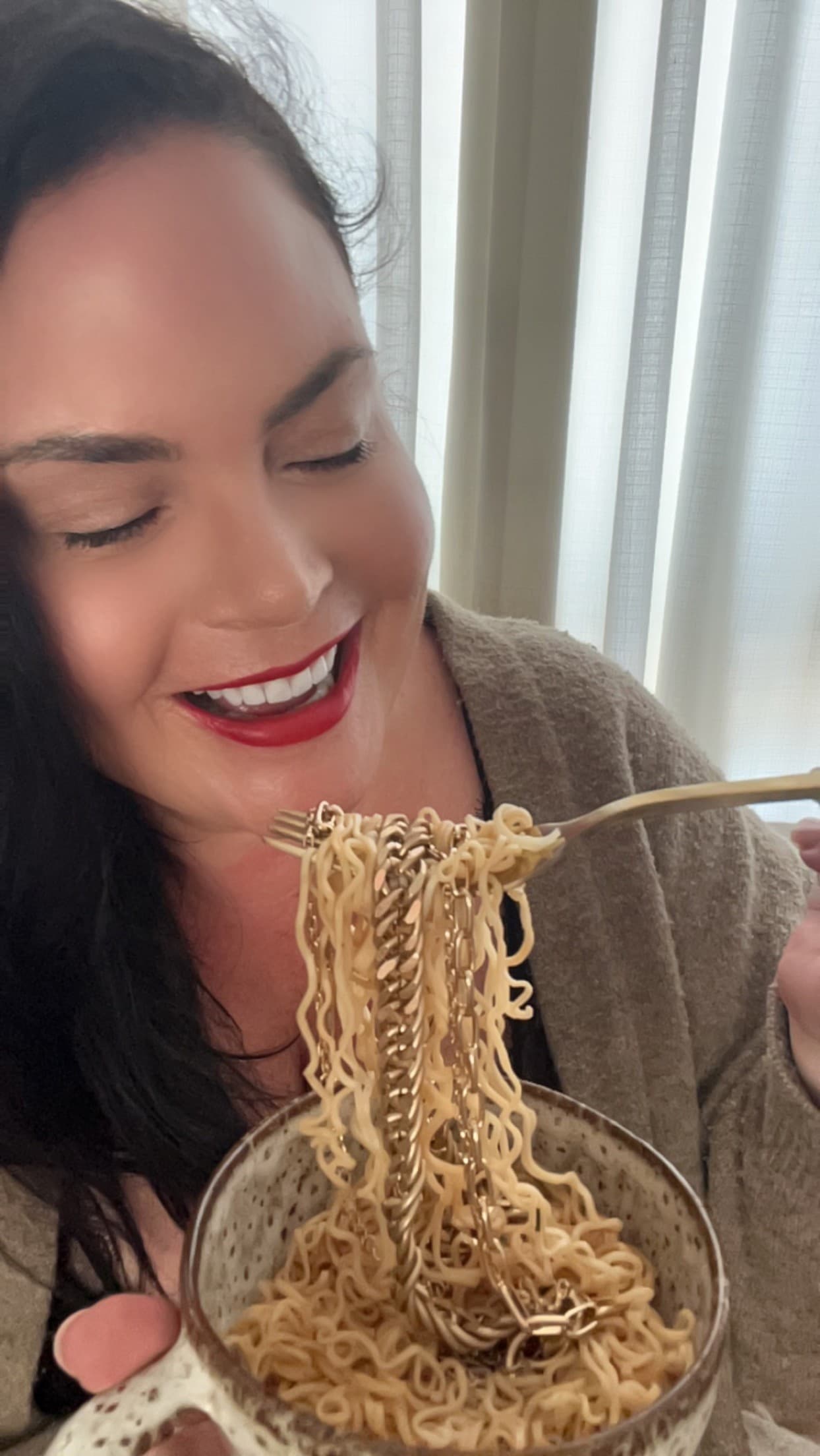 Esther smiling with her eyes closed, lifting a forkful of noodles threaded with gold chain necklaces from a glass bowl.