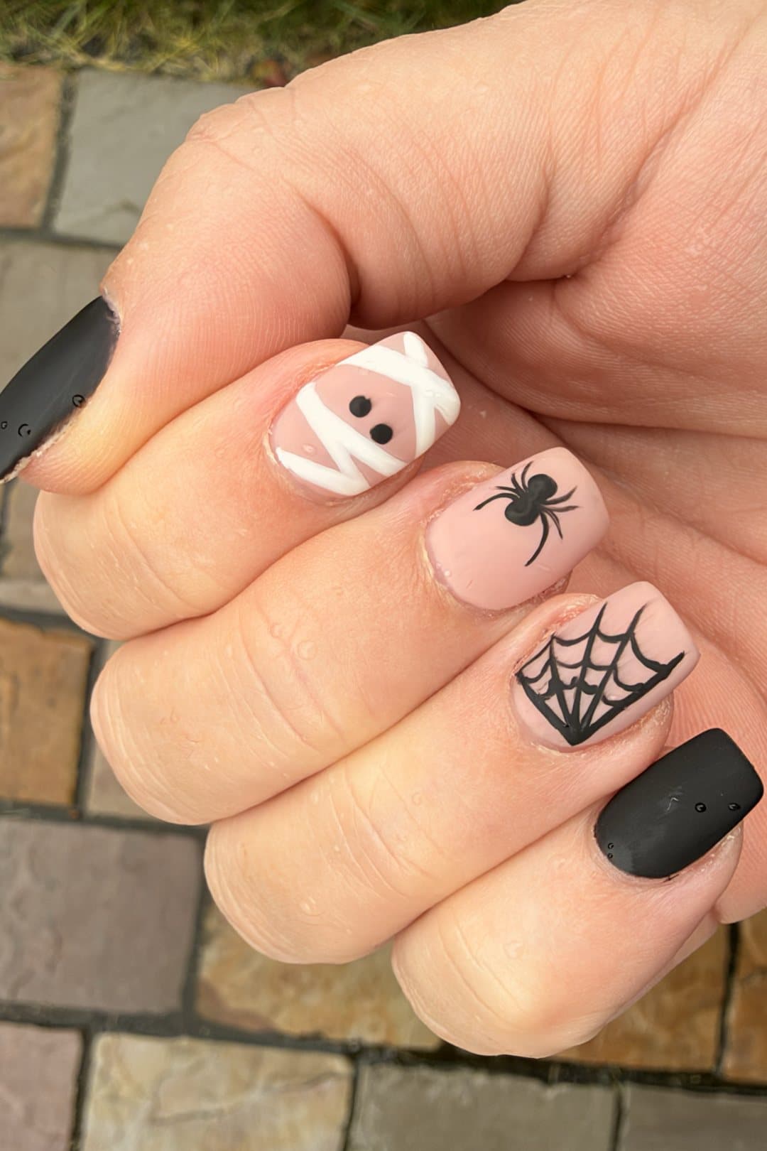 A hand with matte black thumb and little finger nails and three nude accent nails painted with a white-bandage mummy, a black spider and a black spiderweb.