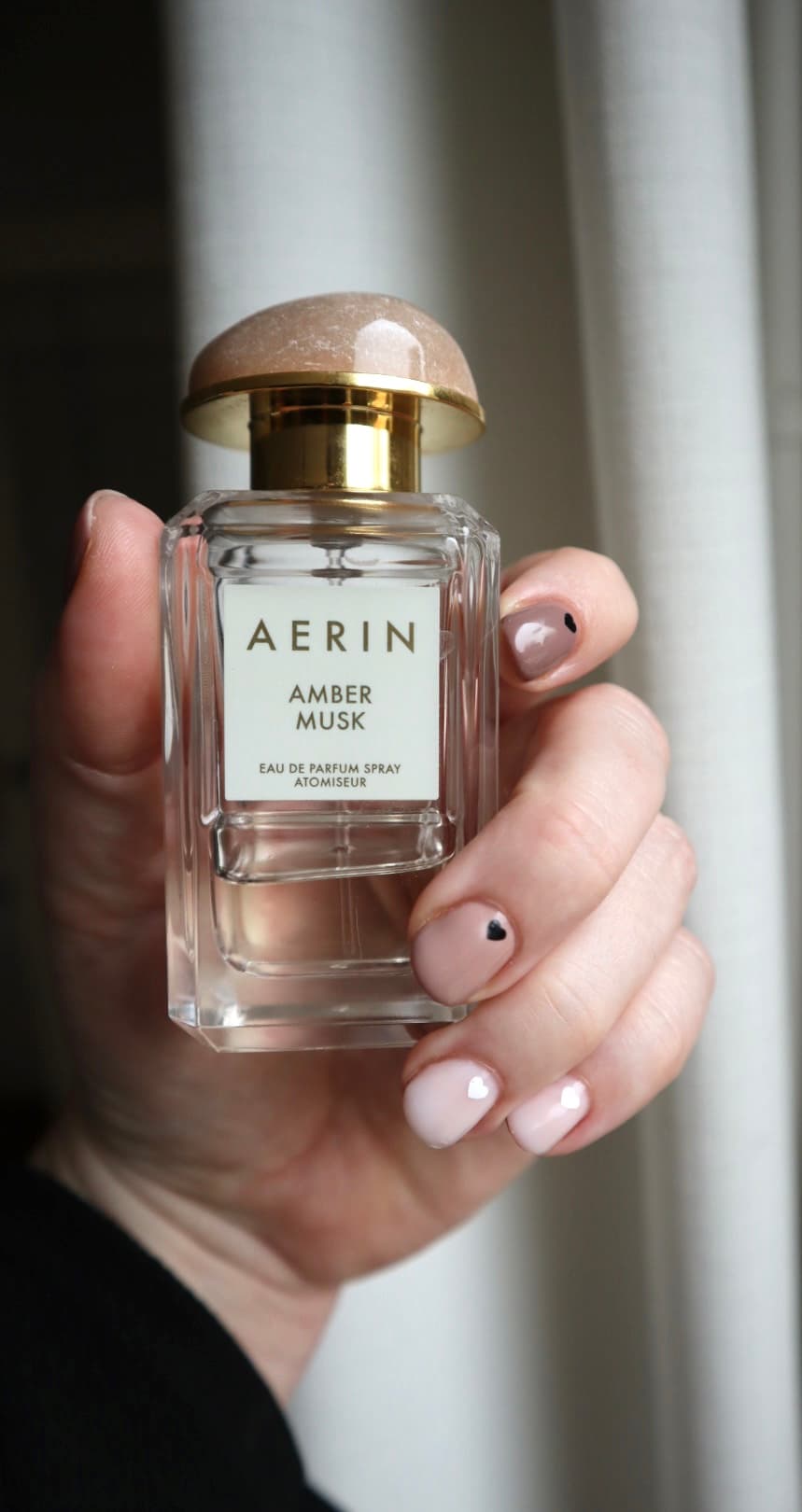 Hand holding a bottle of Aerin Amber Musk perfume in soft natural window light.