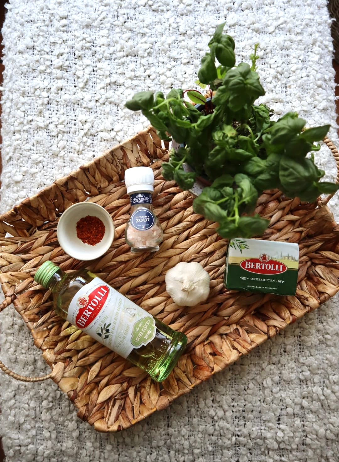 Bertolli olive oil and unsalted butter beside a fresh basil plant, a whole garlic bulb, chilli flakes and Himalayan salt on a woven tray.