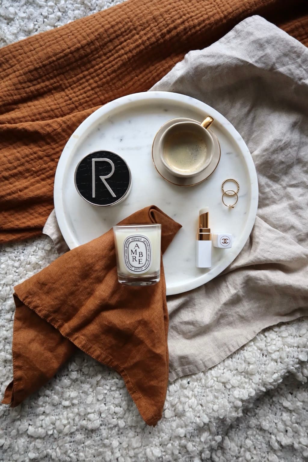 A Diptyque Ambre candle, a cup of coffee on a marble tray, a monogrammed compact, a Chanel lipstick and two gold rings, resting on a rust-coloured linen napkin.