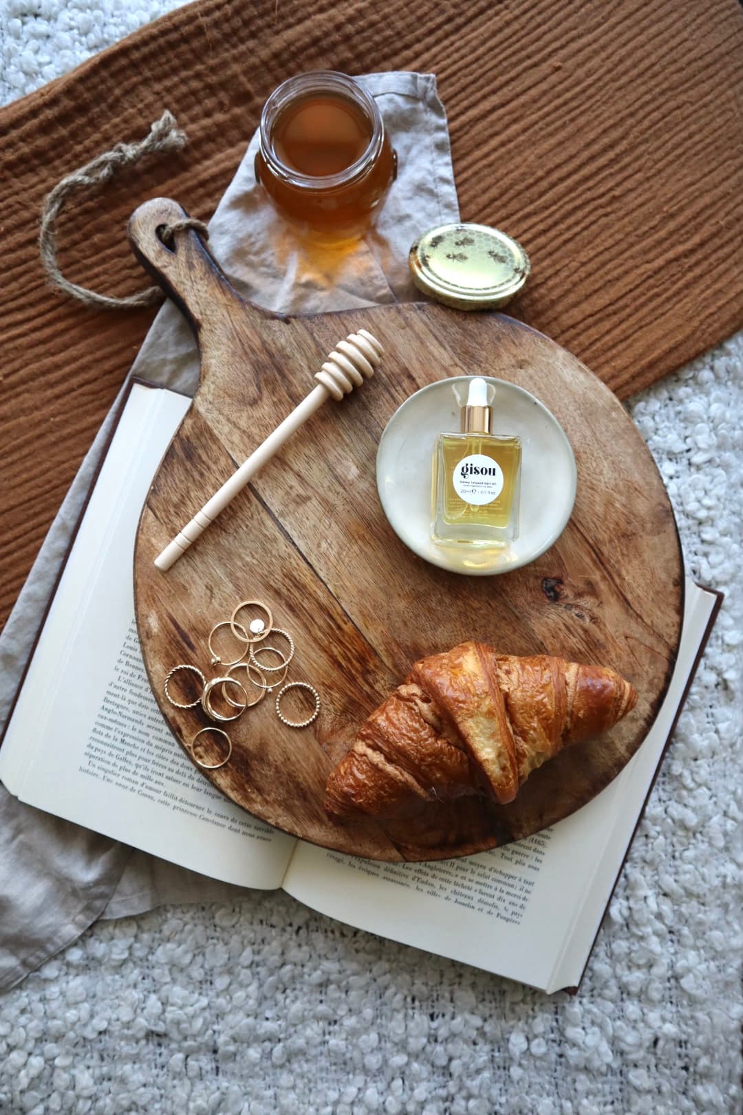 Gisou honey-infused hair oil and a jar of honey beside a croissant and stacked gold rings on a wooden board, resting on an open book.