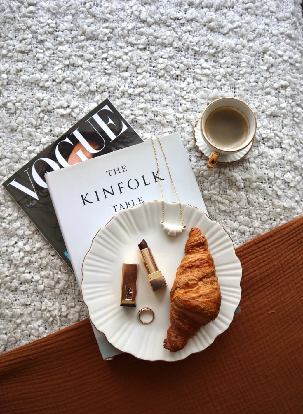 A flatlay of Vogue magazine and The Kinfolk Table book with a gold necklace, a YSL lipstick and compact, a ring, a croissant and a cup of coffee on a rust-coloured cloth.