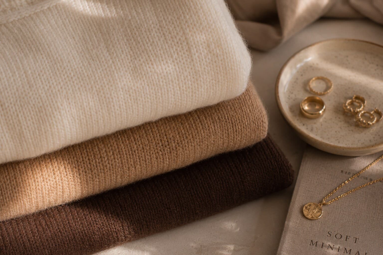 Folded knitwear in cream, camel and chocolate tones arranged beside gold jewellery.