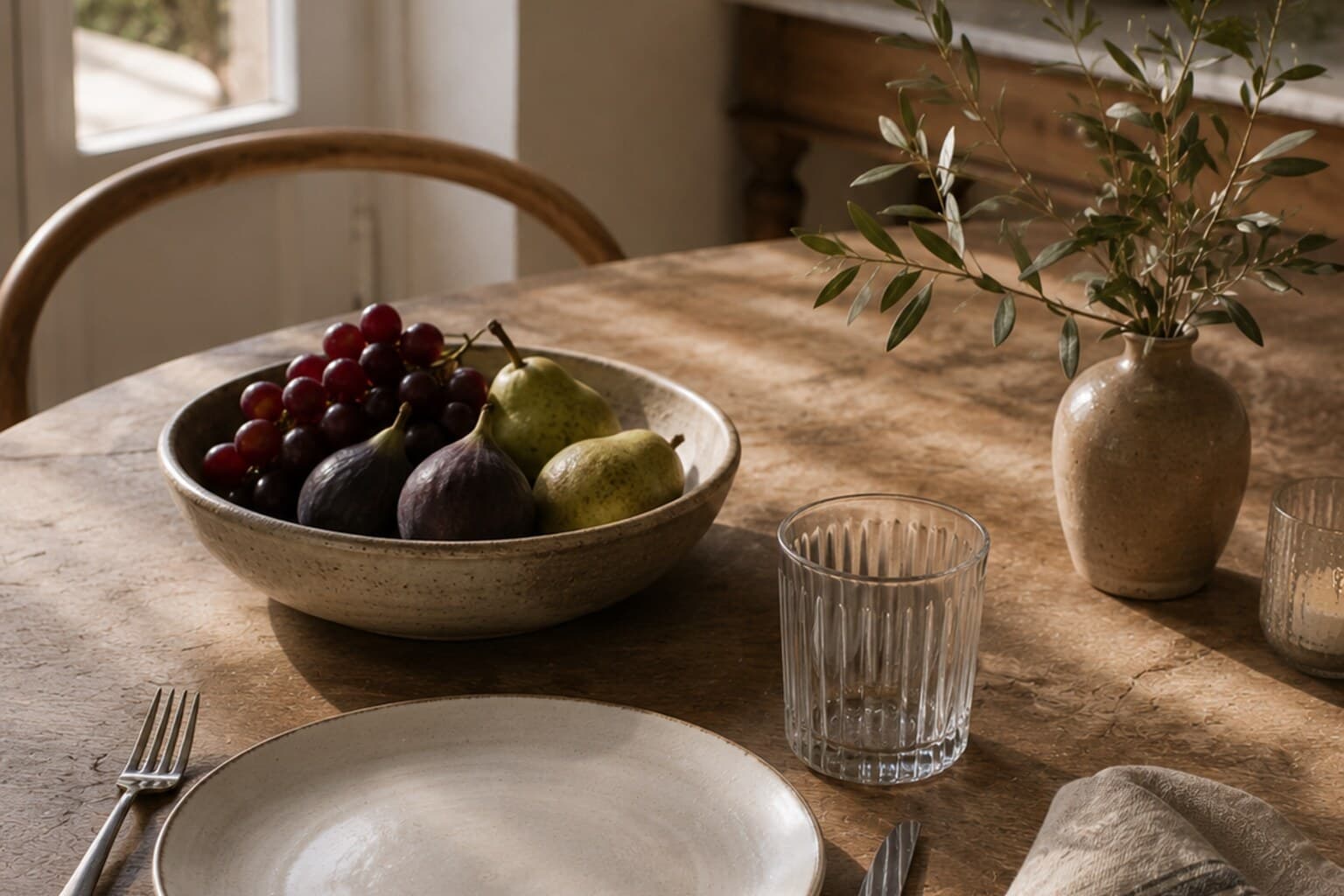 A simply set everyday table with a proper plate, glass, napkin and a low bowl of fruit.