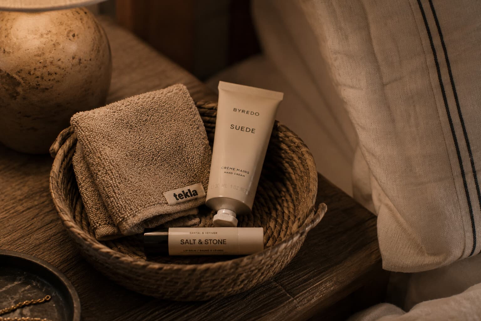 A small, simple care basket with a face cloth, lip balm and hand cream beside a bed.