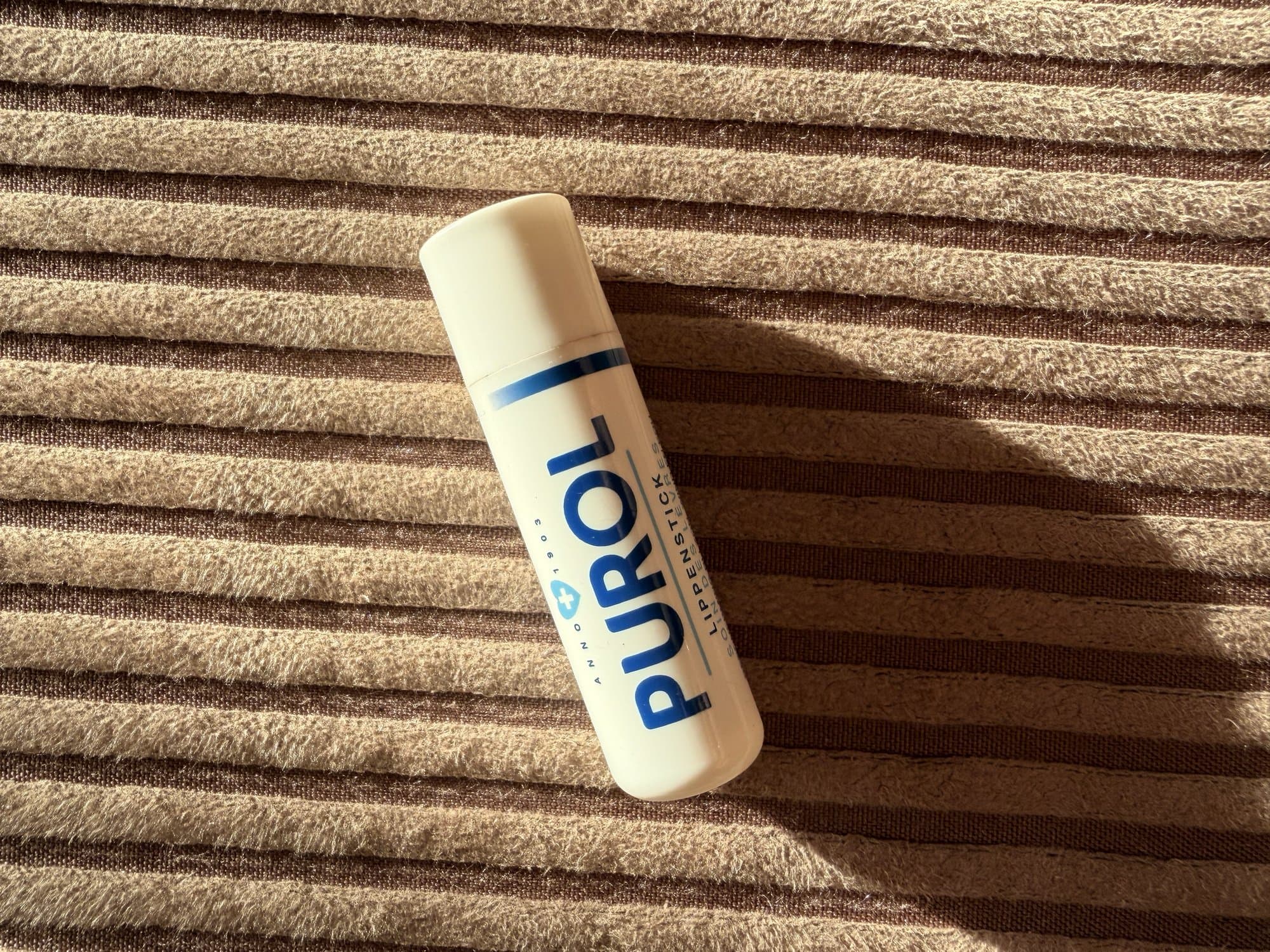Purol lip stick lying on a ribbed brown fabric in warm sunlight.