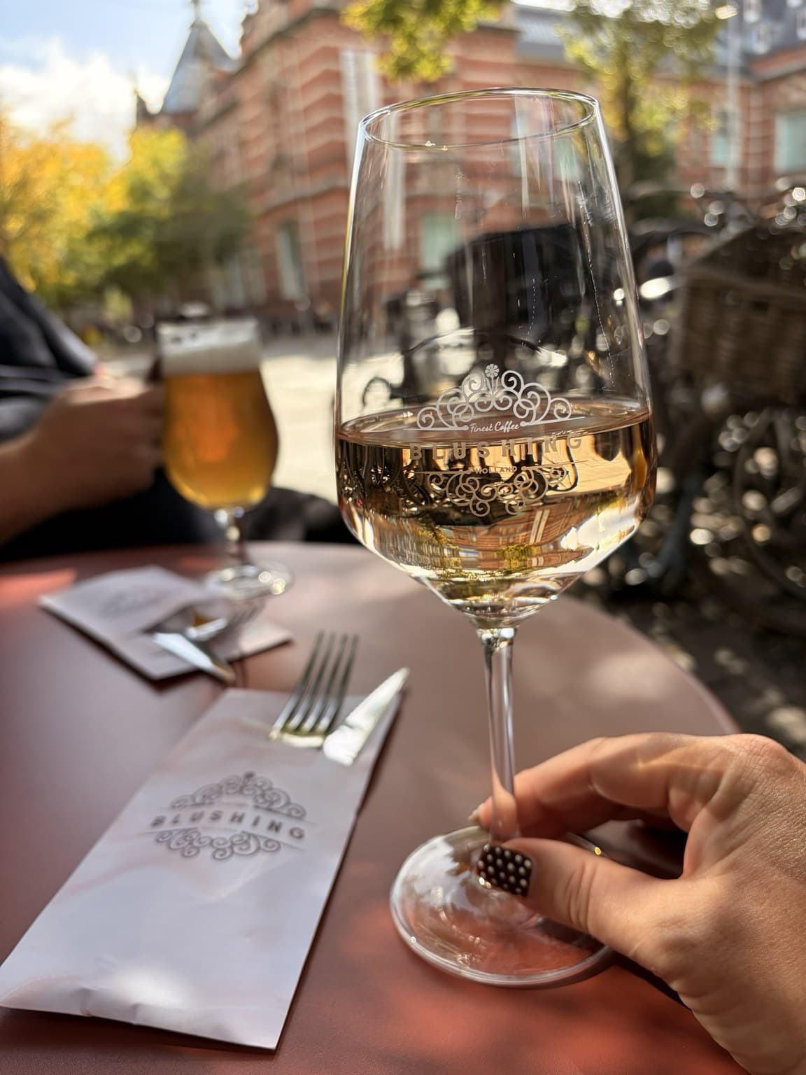 A glass of white wine on a sunny terrace table, with a beer and a grand red-brick building in the background