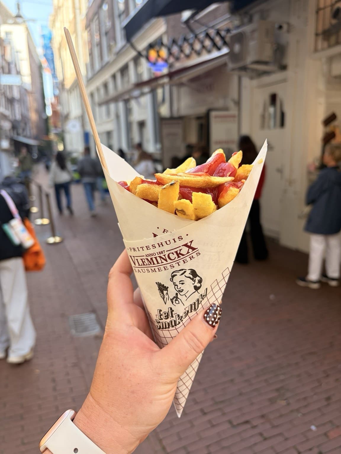 A hand holding a paper cone of Vleminckx Sausmeesters fries with ketchup, on a busy Amsterdam street