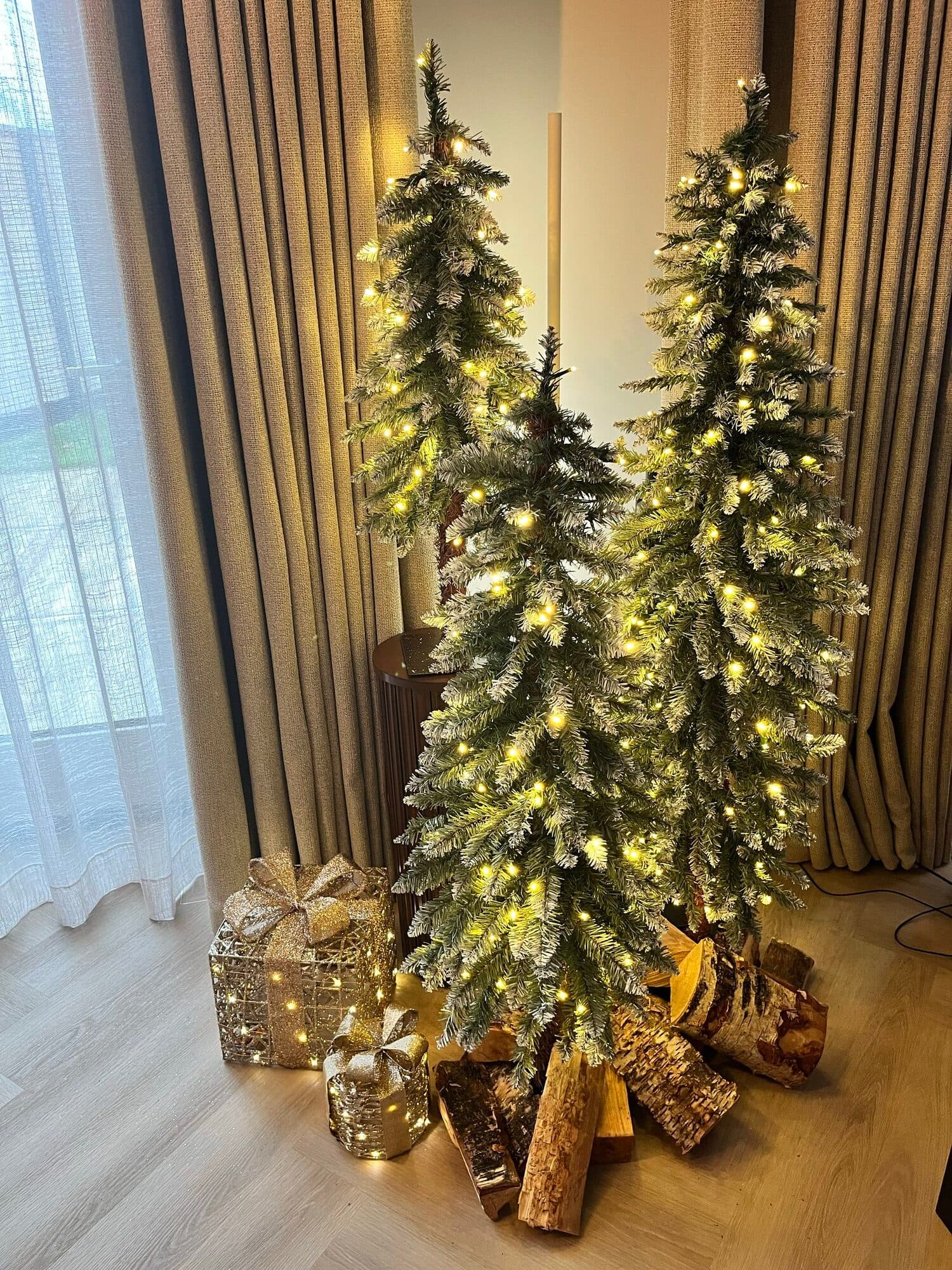 A flocked, lit Christmas tree at home surrounded by wrapped gifts and stacked birch logs