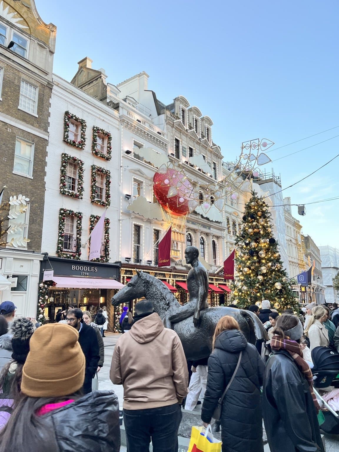 A busy Bond Street on a bright winter afternoon, shopfronts dressed with red baubles and a Christmas tree in the distance.