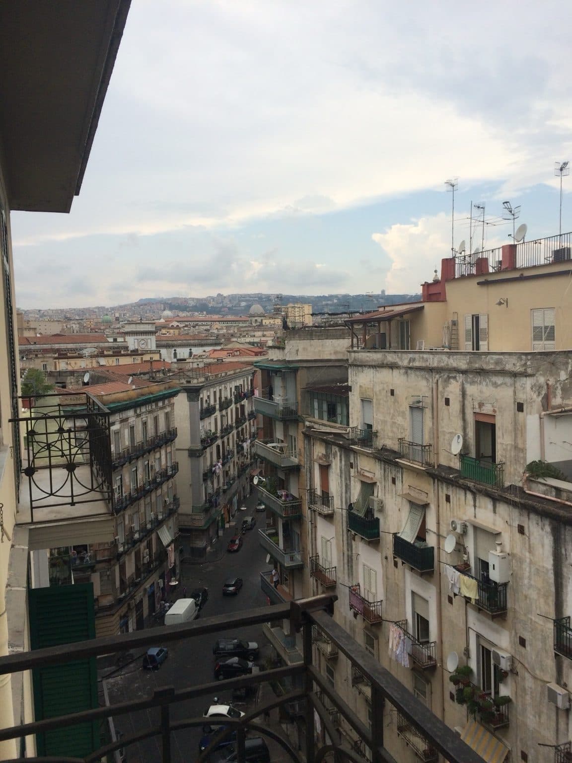A view down a narrow Naples street from a balcony, with dense rooftops and a hazy hillside beyond under a cloudy sky