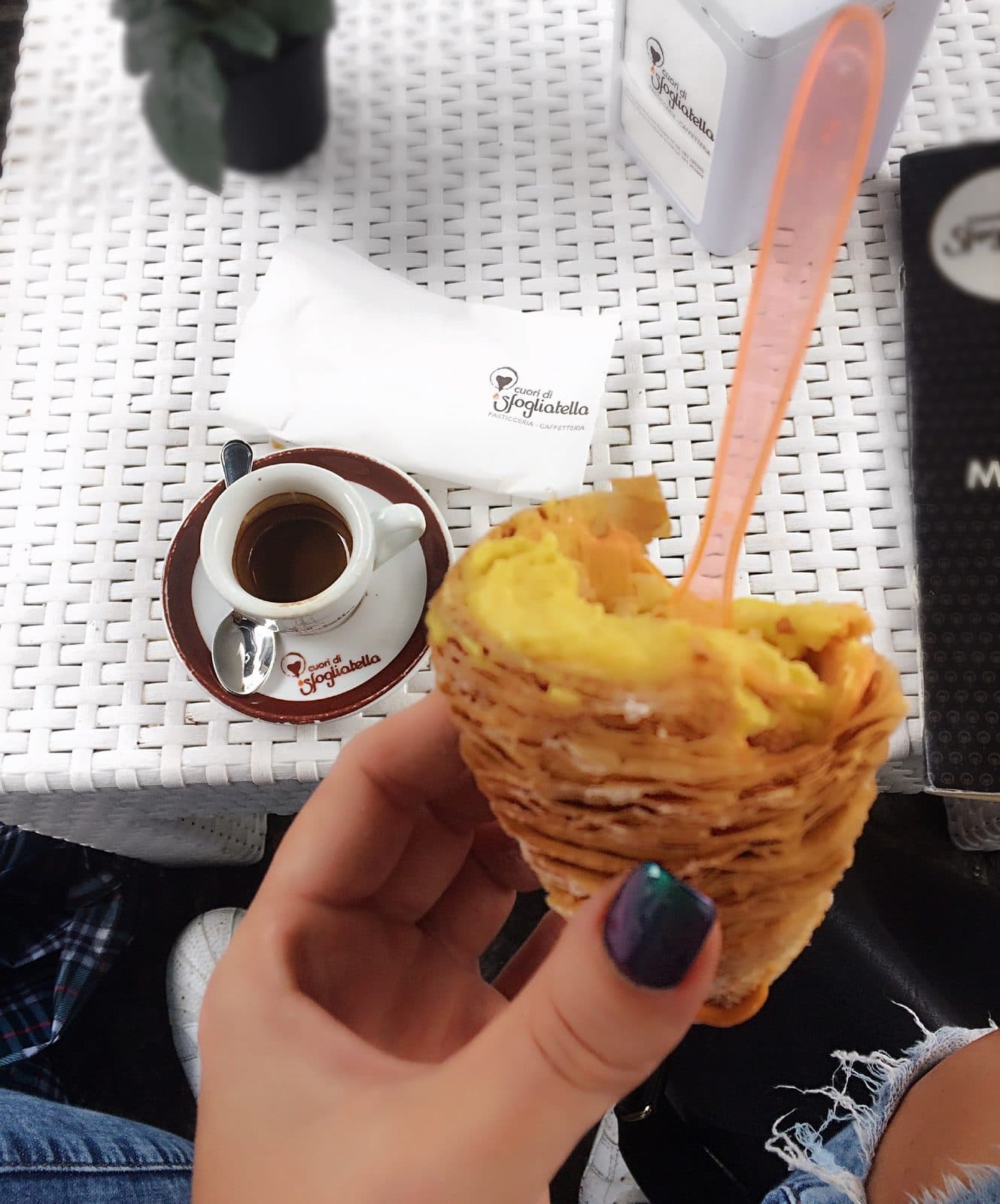 A hand holding a sfogliatella pastry with crisp layered shell beside a tiny espresso cup on a white table
