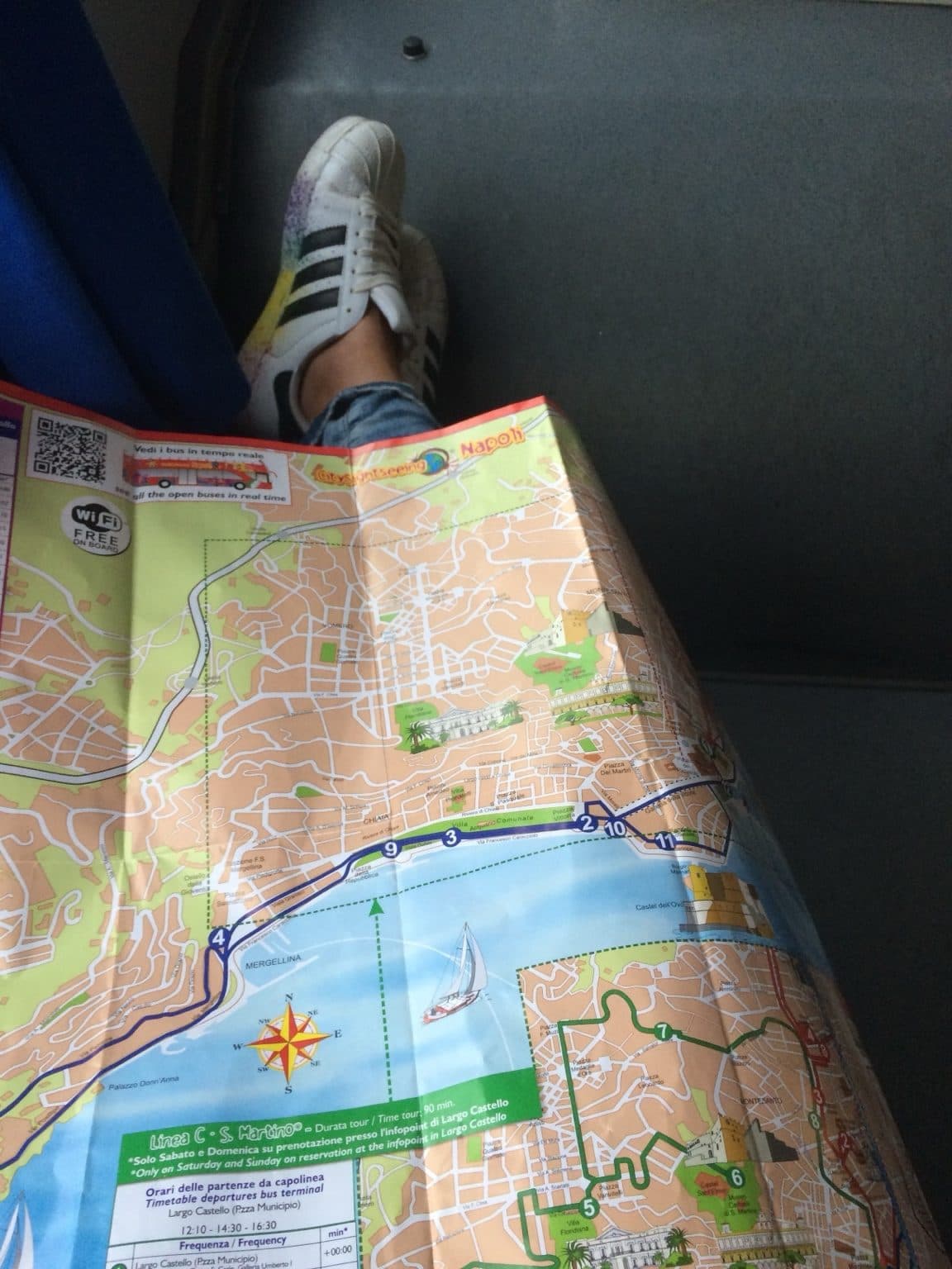 A paper City Sightseeing map of Naples unfolded over a lap, with white trainers at the top of the frame
