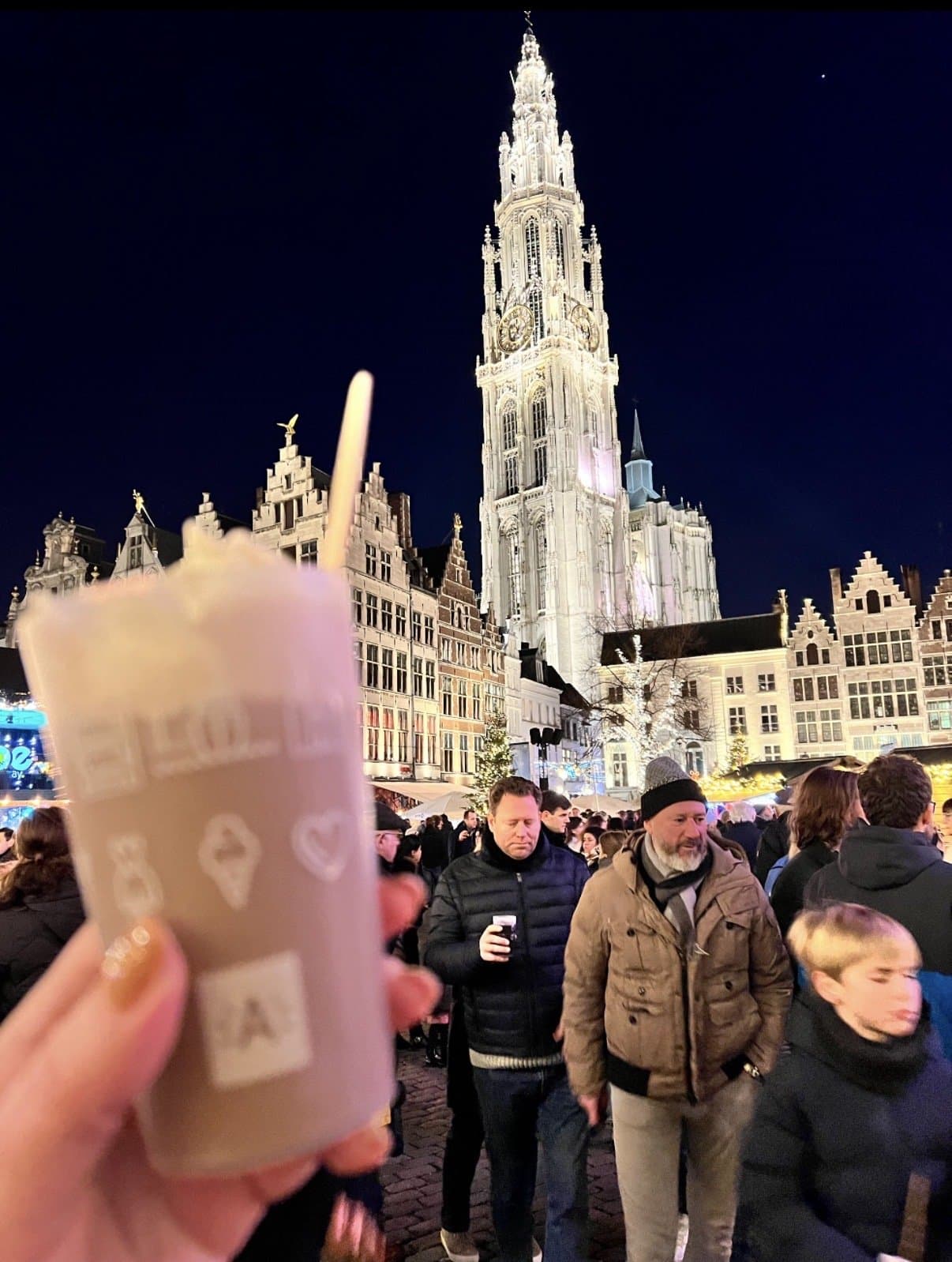 A drink held up at night with the illuminated tower of Antwerp's Cathedral of Our Lady behind.