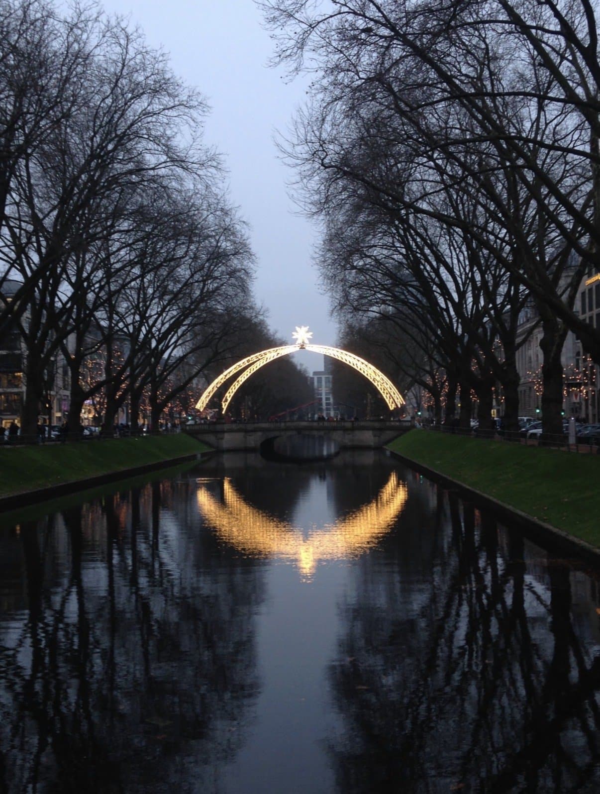 An illuminated arch of festive lights reflected in still canal water at dusk.
