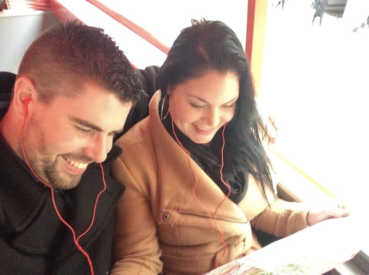Esther and Bas sitting together on an open-top sightseeing bus, both wearing audio-guide headphones.
