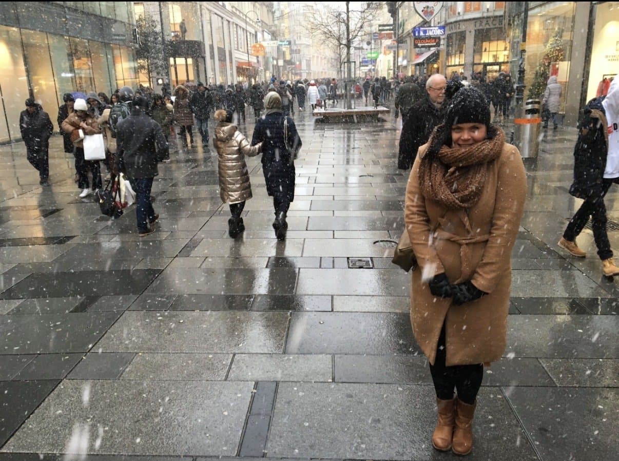 Esther smiling in a camel coat and scarf on a snowy pedestrian shopping street as snow falls.