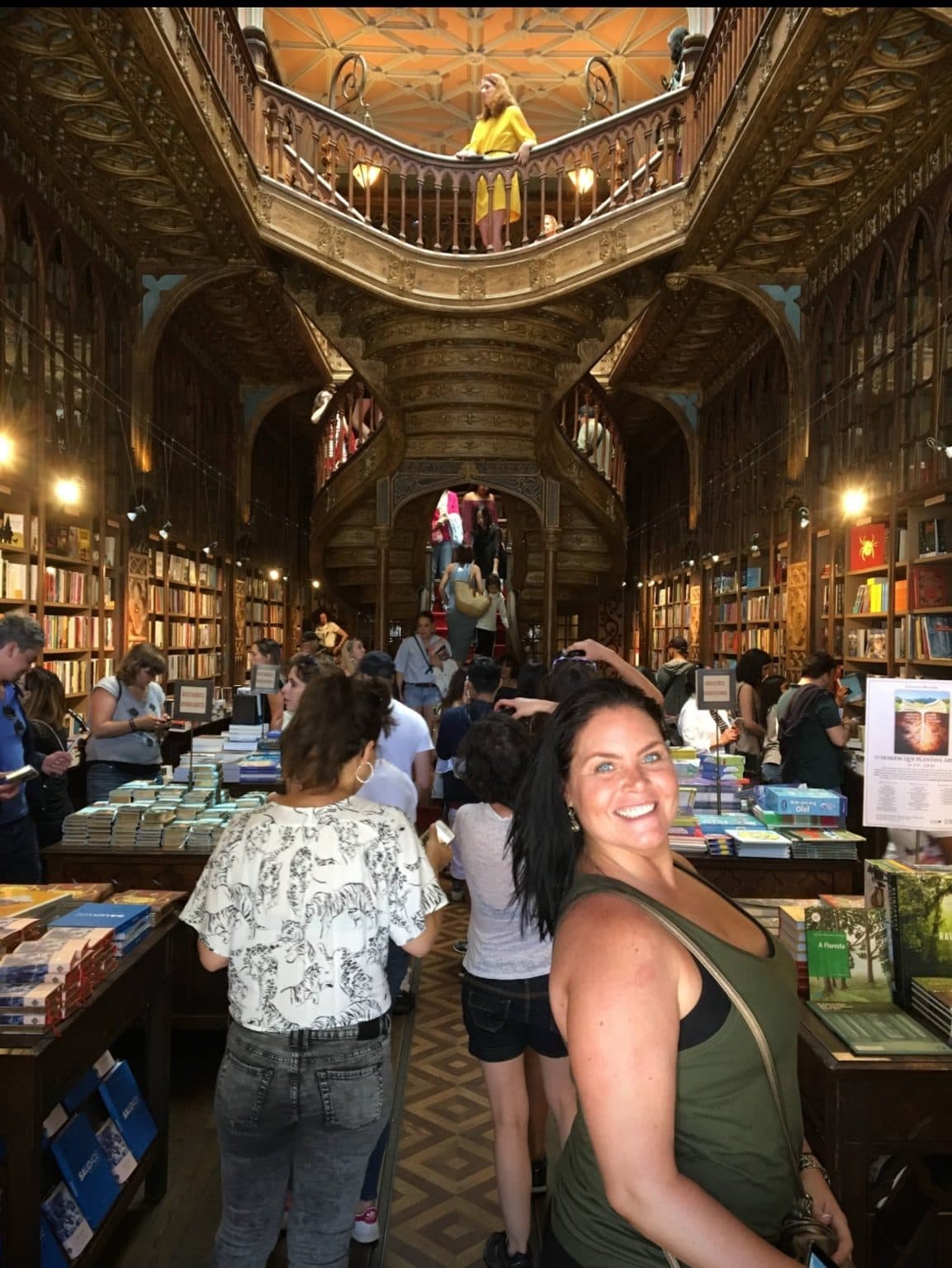 Esther smiling in the crowded main hall of Livraria Lello in Porto, with its carved wooden staircase and book-lined shelves behind her