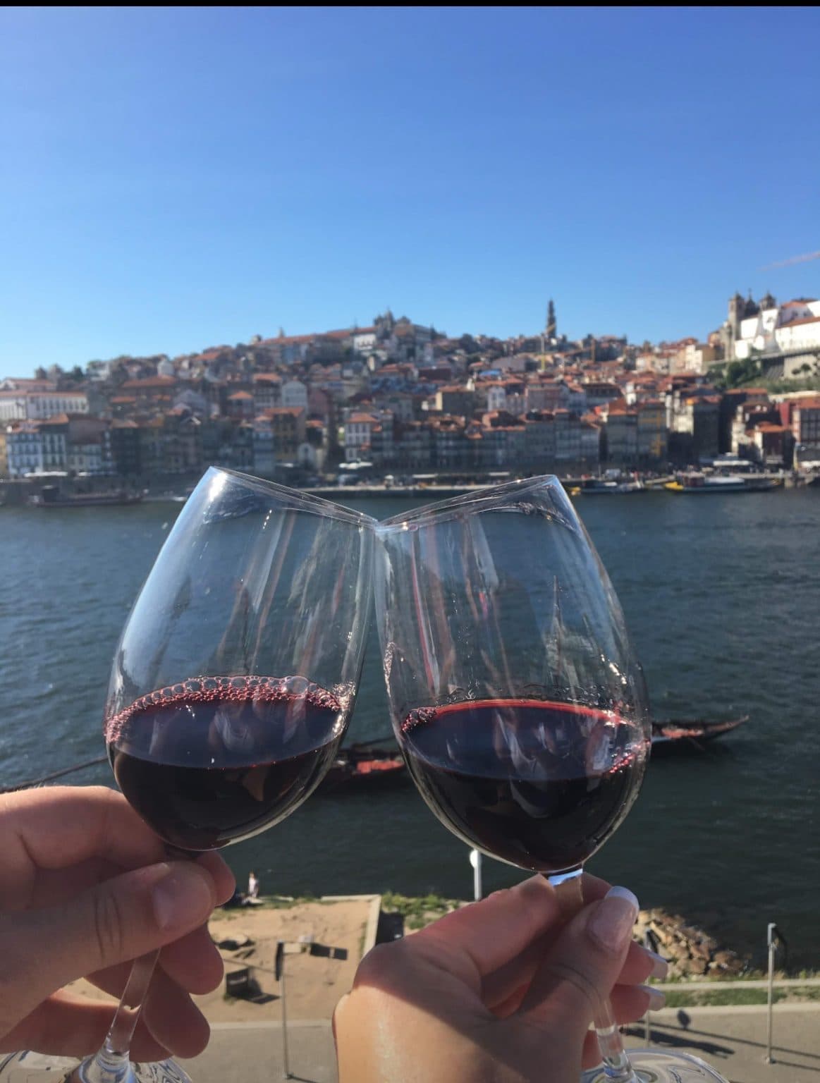Two hands clinking glasses of red wine in front of the Douro river, with Porto’s stacked hillside under a bright blue sky