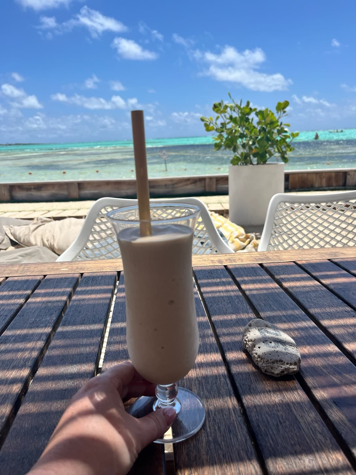 Cream-coloured iced drink held above a wooden table with turquoise sea in the background