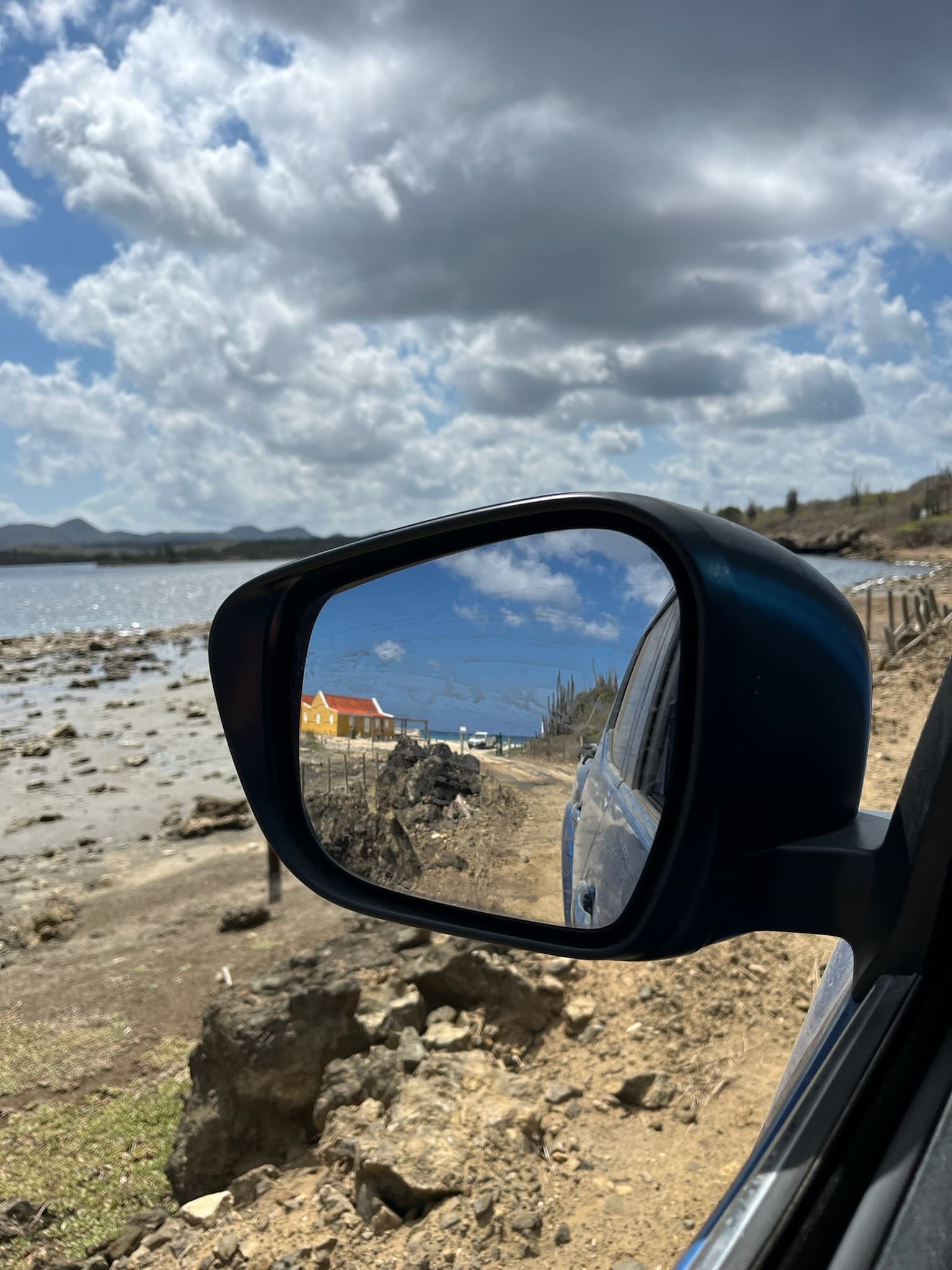 Coastal road and a yellow house reflected in a car side mirror under a cloud-filled sky