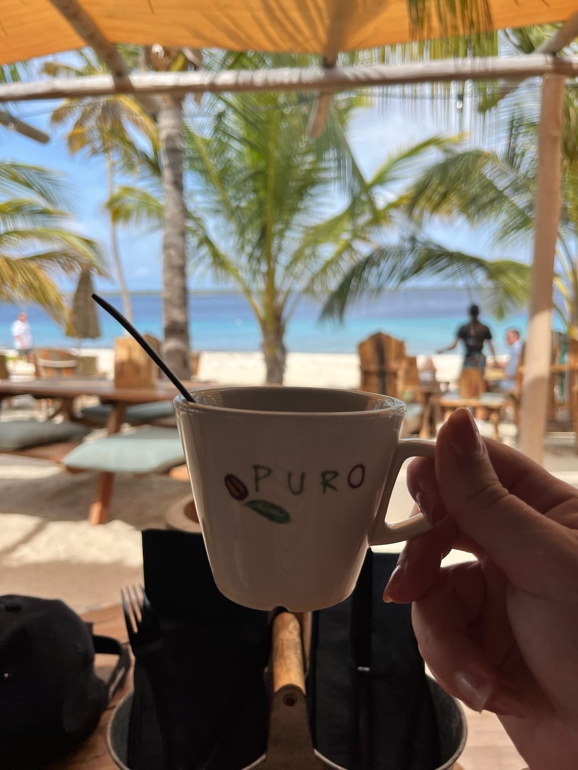 Hand holding a coffee cup at a shaded beach restaurant with palms and blue sea behind it