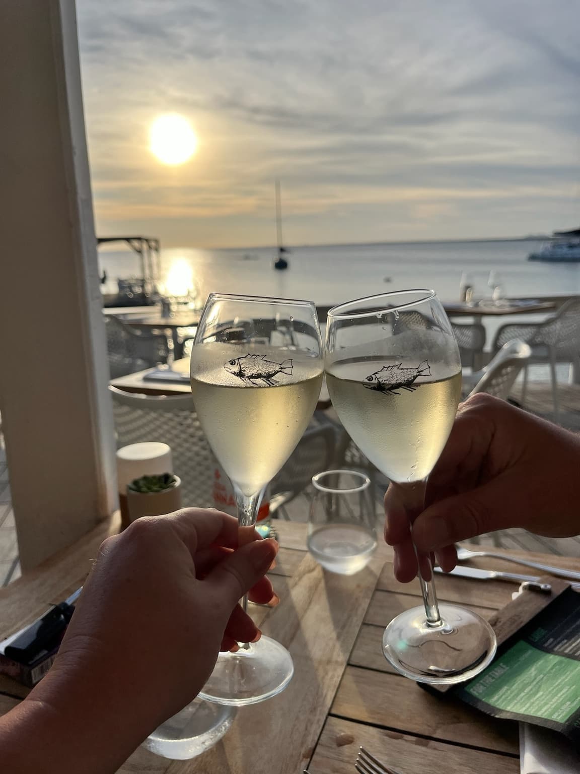 Two hands raising glasses of chilled white wine at a waterfront table near sunset