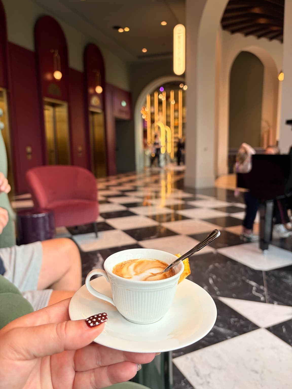 A white coffee cup held in the Efteling Grand Hotel foyer, with patterned flooring and golden lifts behind it