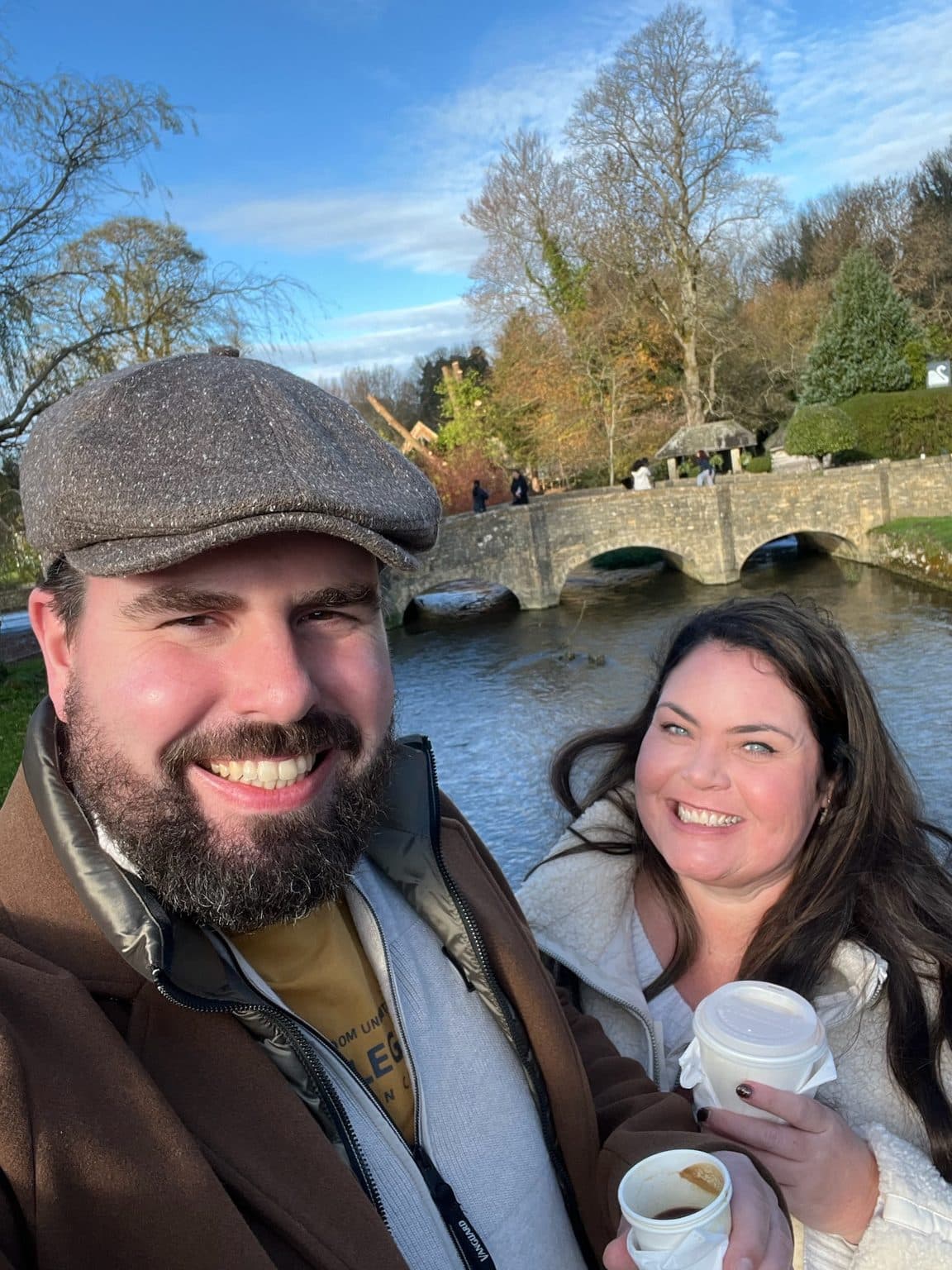 Esther and Bas smiling with takeaway coffees beside the river and old stone bridge in Bibury