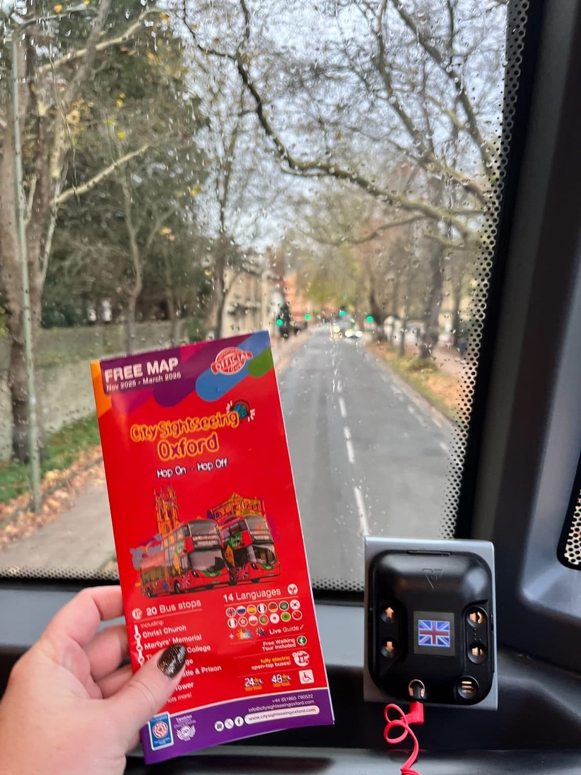 A red City Sightseeing Oxford hop-on hop-off map held up inside the bus, with rain on the windscreen and a tree-lined road ahead