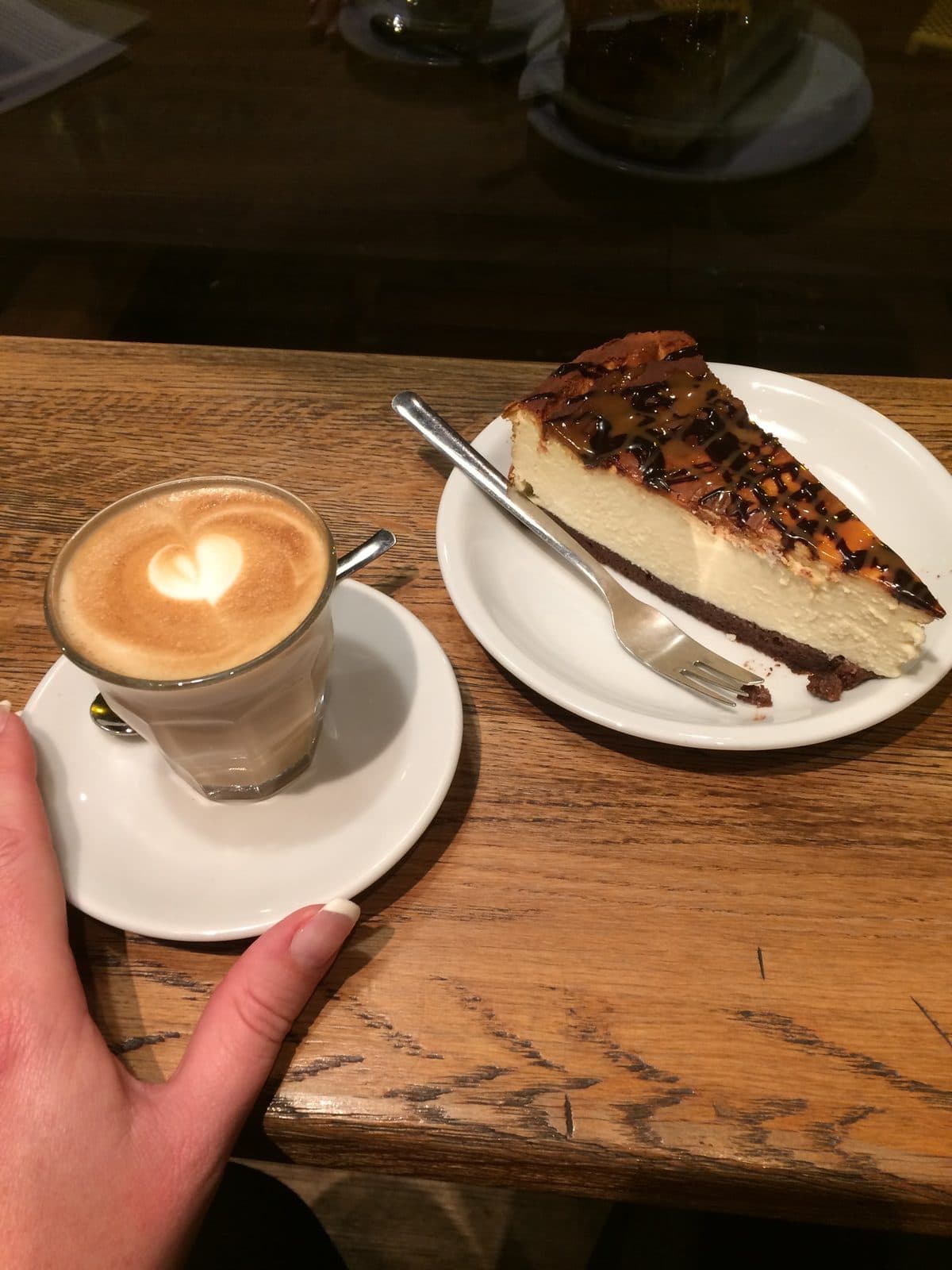 A cappuccino with latte art beside a slice of cheesecake on a wooden café table, a hand resting at the edge