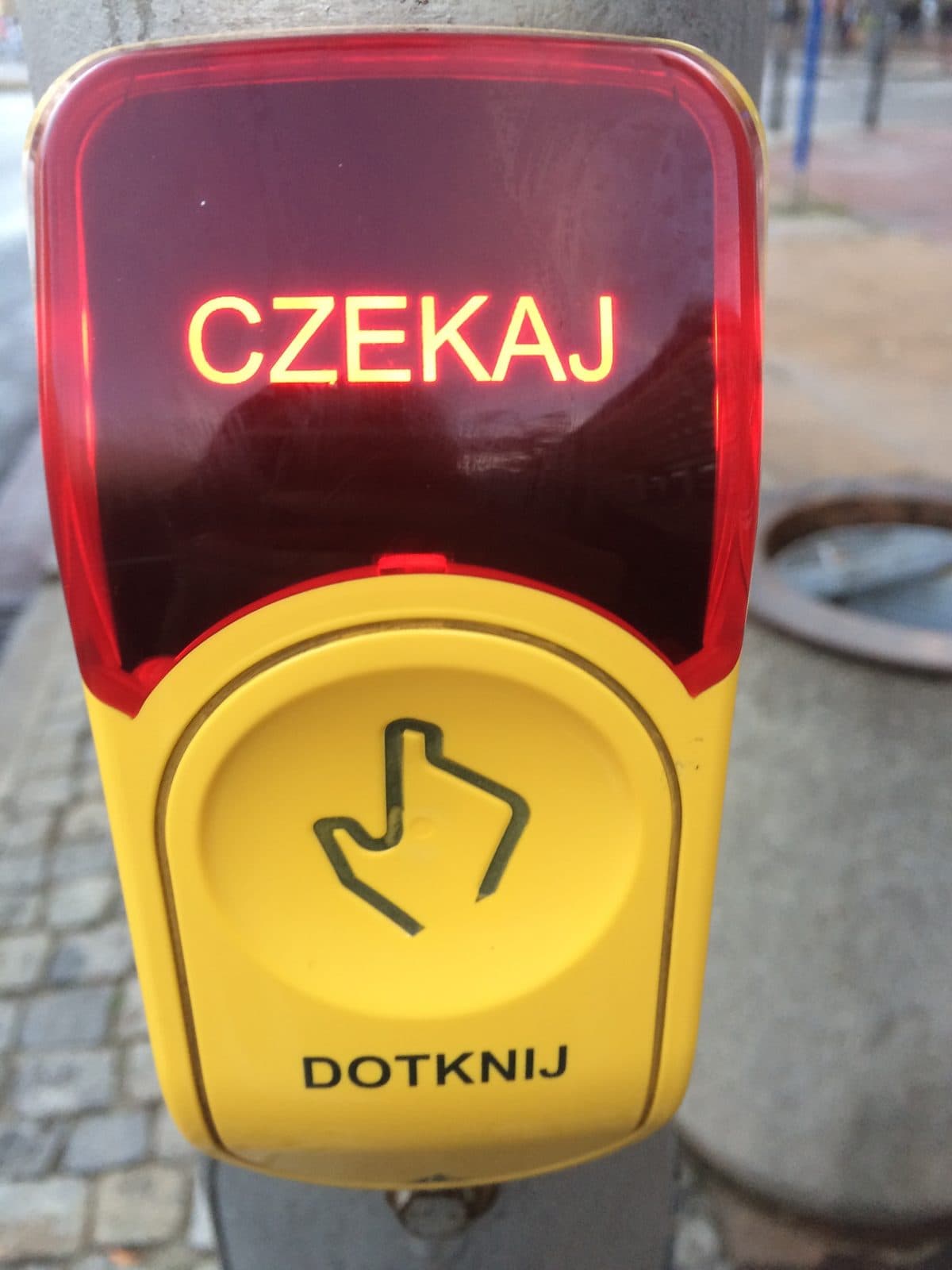 A red-and-yellow pedestrian crossing button reading “CZEKAJ” on a Warsaw street