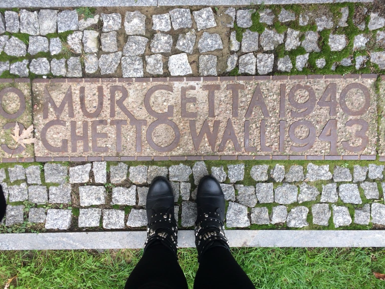 Black boots standing on grass in front of a pavement marker reading “Mur Getta 1940, Ghetto Wall 1943”