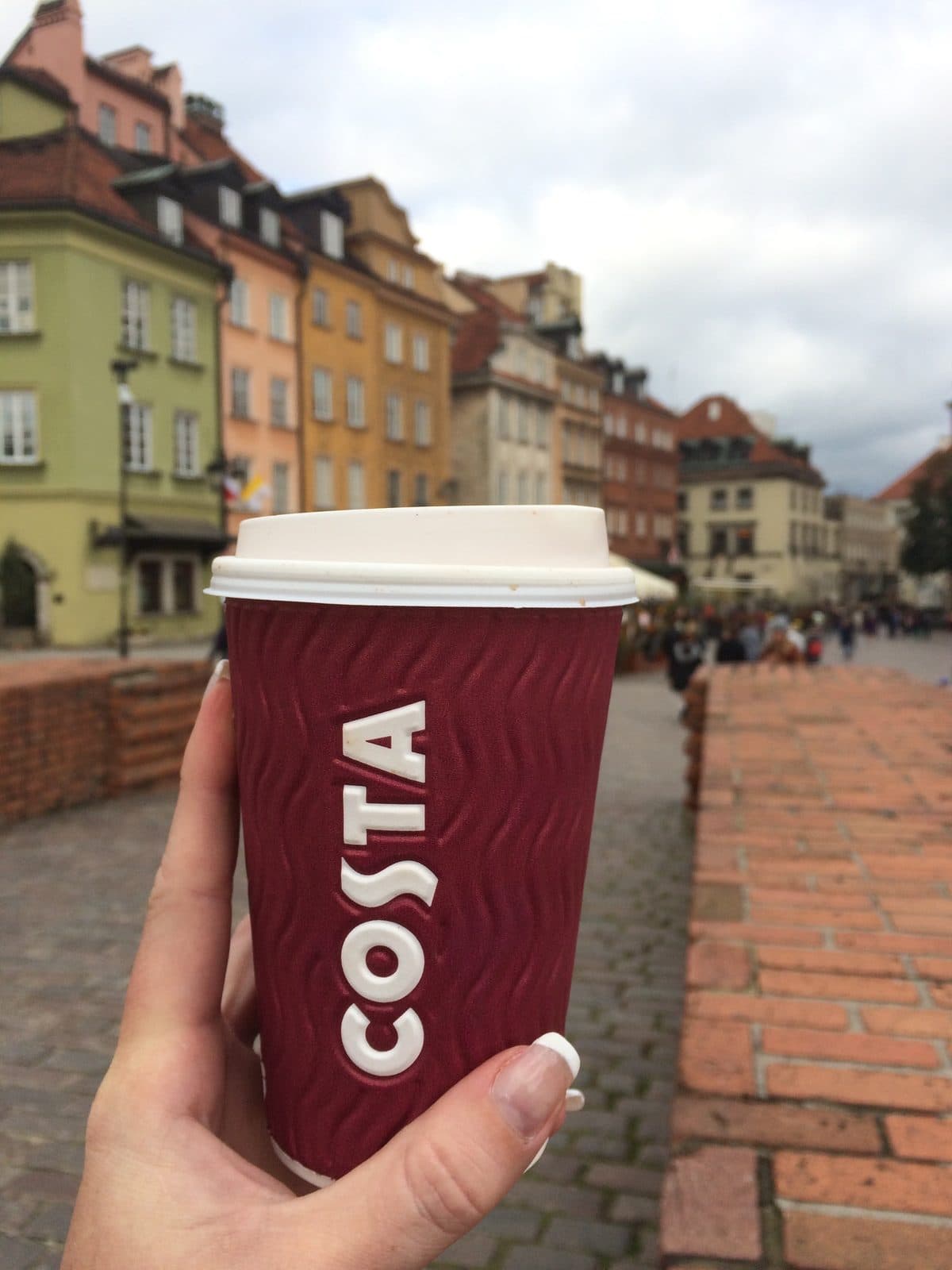 A hand holding a takeaway coffee cup in front of the colourful historic buildings of Warsaw’s Old Town on a cloudy day