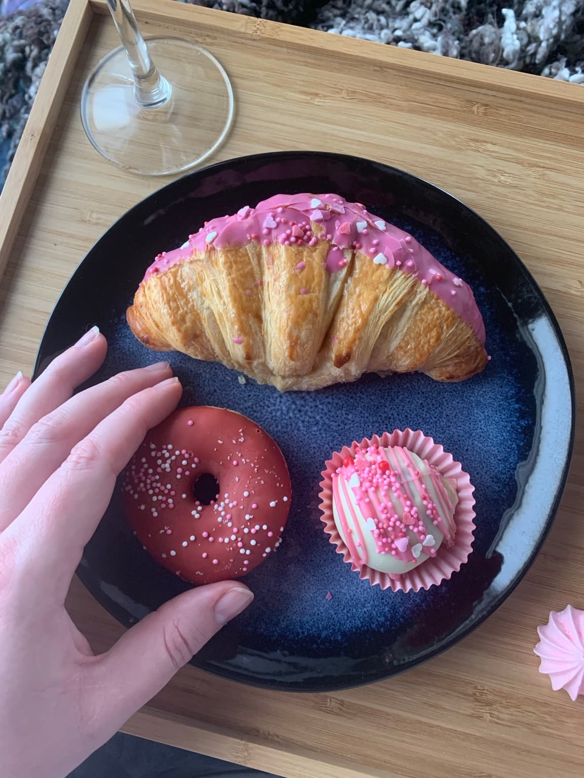 A pink-decorated croissant, doughnut and small sweet treat on a dark plate on a wooden tray.